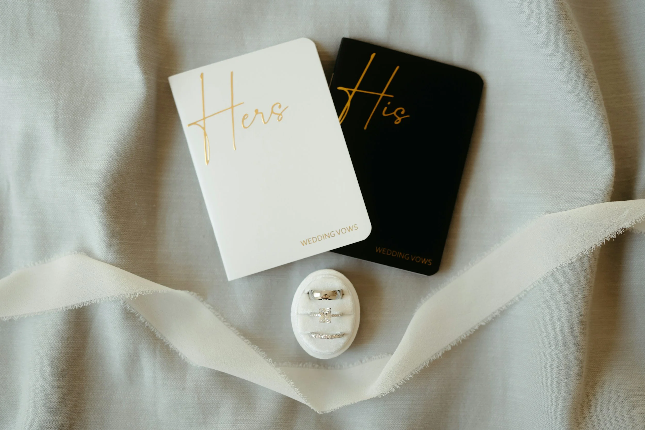 Two wedding vow books, one white and one black, with gold lettering that says 'Hers' and 'His,' along with a set of rings on a white ring box, arranged on a light fabric background with a white ribbon.