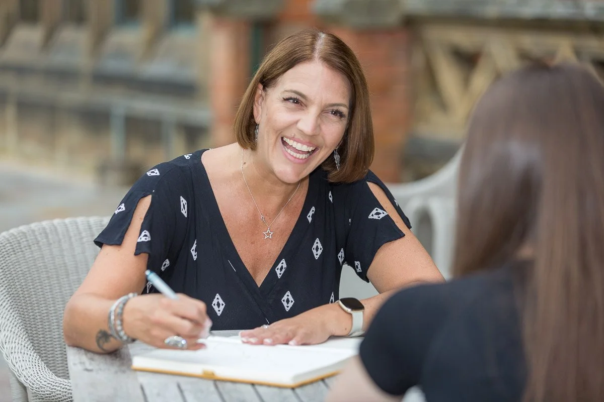A woman with short brown hair, wearing a black top with cut-out shoulders, smiling and talking to another woman with long brown hair, outdoors in a casual setting.