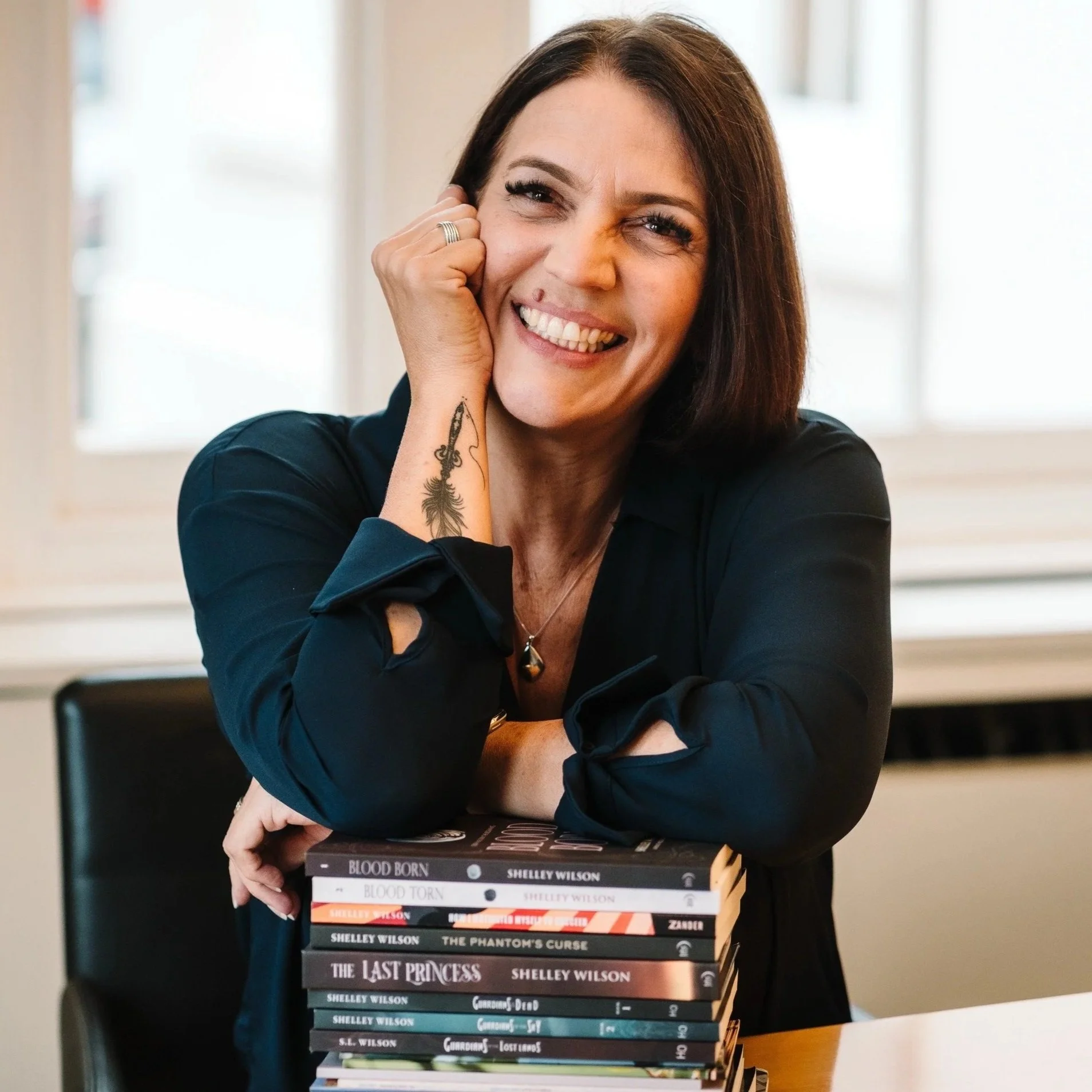 A woman with dark hair, smiling and resting her face on her hand, leans on a stack of books on a table.