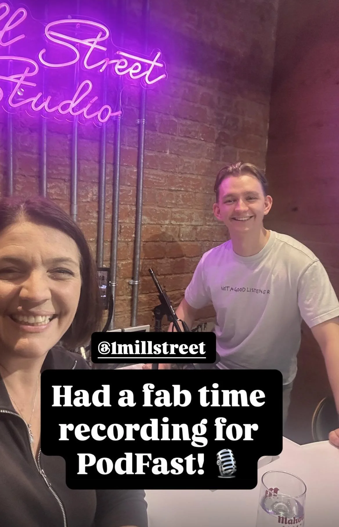 smiling women and a man sitting at a table in a recording studio with a neon sign that reads 'Street Studio' in pink and purple. The woman on the left has shoulder-length dark hair, and the man wears a white t-shirt. There is a microphone and a glass