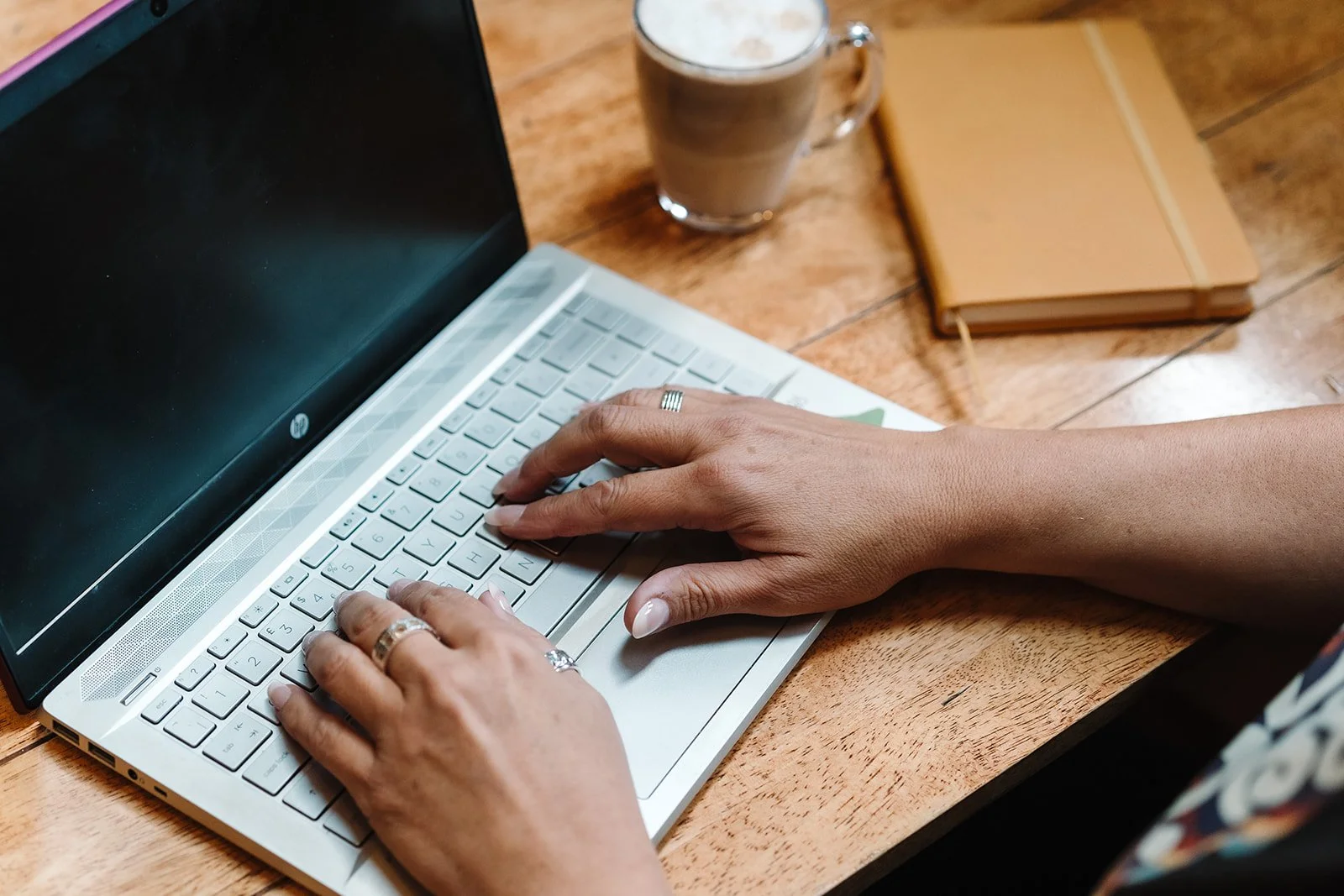 Person typing on a silver laptop keyboard at a wooden table, with a coffee drink, a closed notebook, and a pen nearby.