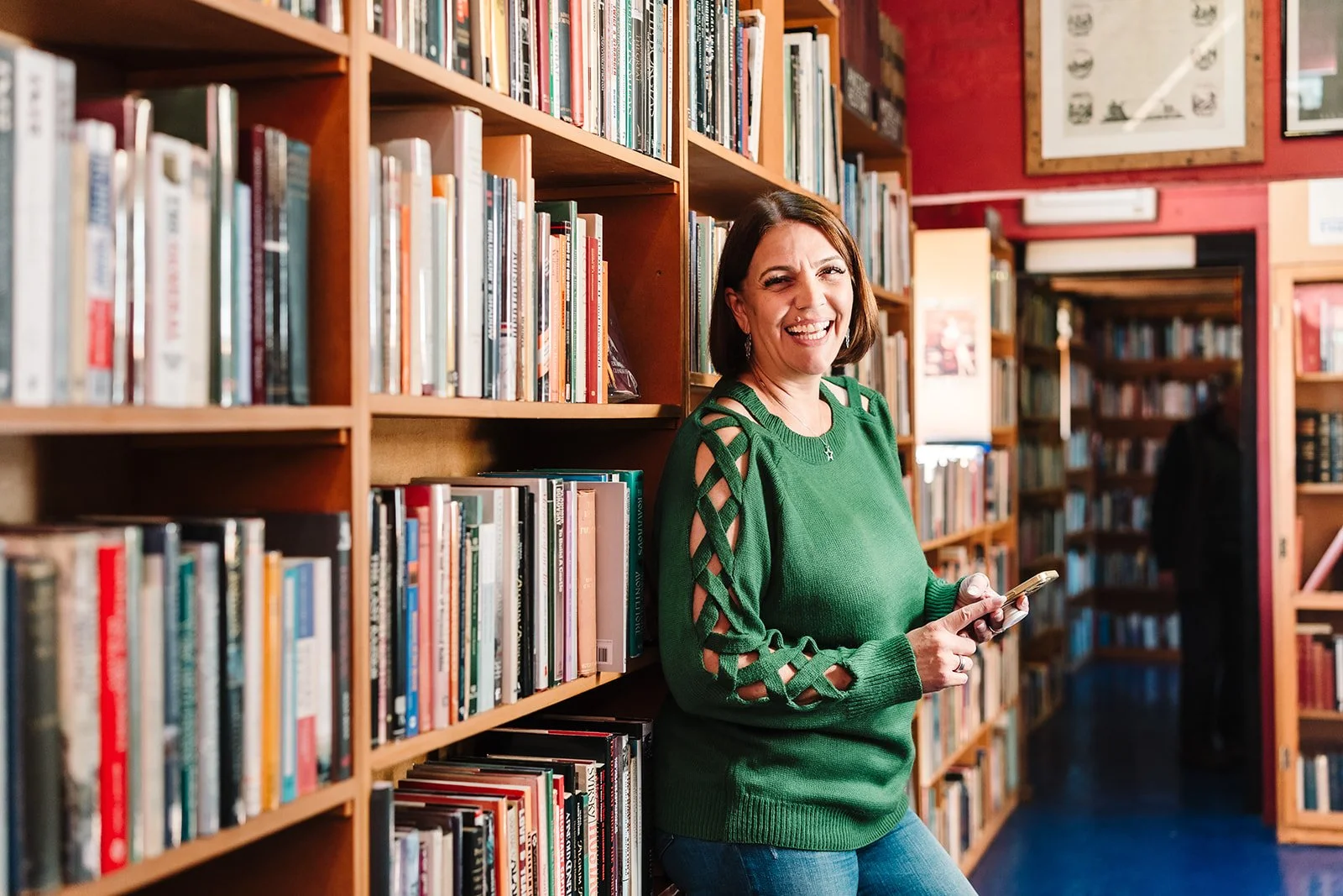 A smiling woman in a green sweater with cut-out shoulder details standing in a bookstore or library, leaning against a wooden bookshelf filled with books, holding a smartphone.