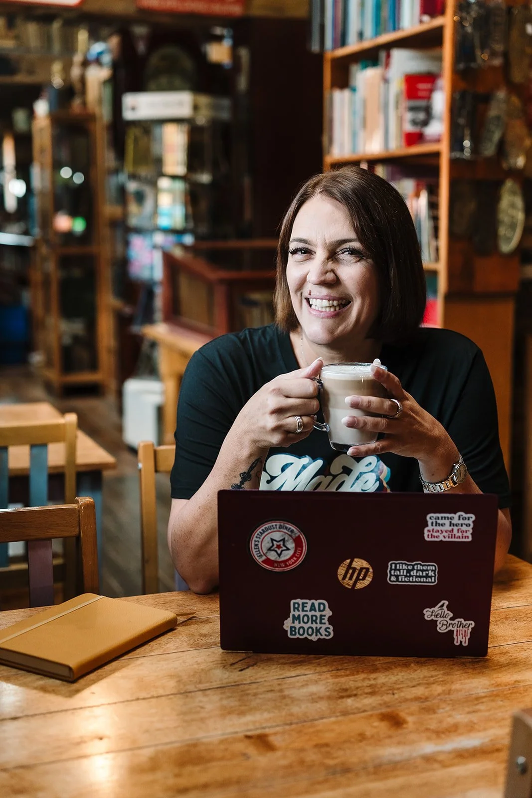 A woman with short brown hair sitting at a wooden table in a cozy café, smiling and holding a mug of coffee or hot chocolate. There is a closed notebook on the table and a laptop with various stickers in front of her. The background has bookshelves filled with books.