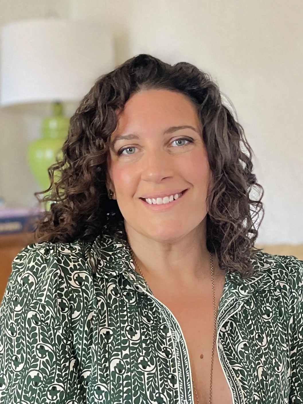 A woman with curly dark hair, wearing a green patterned top, smiling in a room with a green lamp and a beige wall in the background.