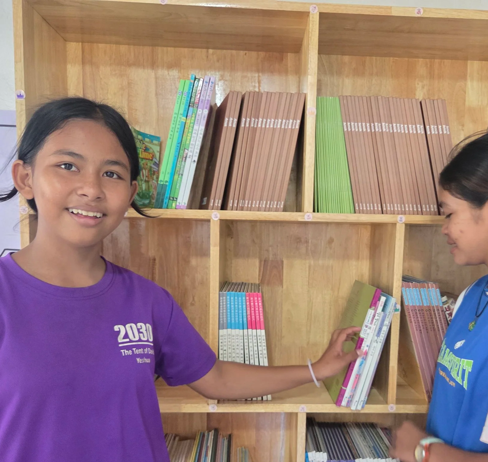 A girl in a purple T-shirt smiling and holding a book in front of a wooden bookshelf, with another girl in a blue T-shirt looking at books on the shelf.