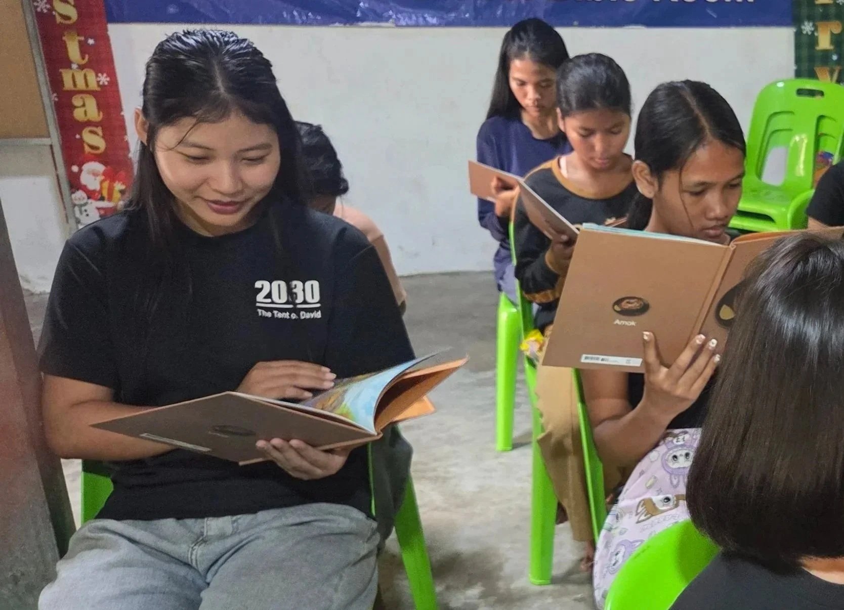 Several girls sitting on green plastic chairs, reading books in a classroom environment.
