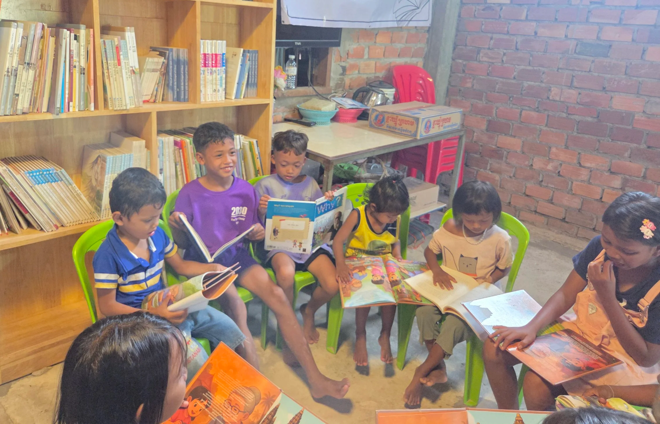 Young children sitting on green chairs reading books in a room with brick walls, a wooden bookshelf filled with books behind them, and a table with a cardboard box and some bowls on it.