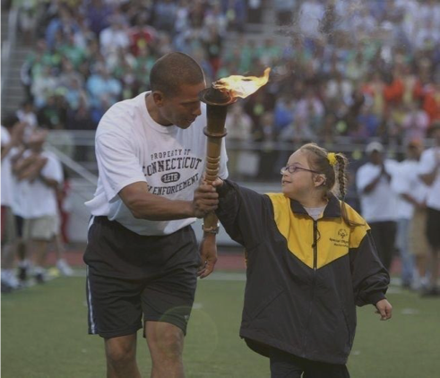 A man and young girl holding a torch with fire during a sports event on a field with a crowd in the background.