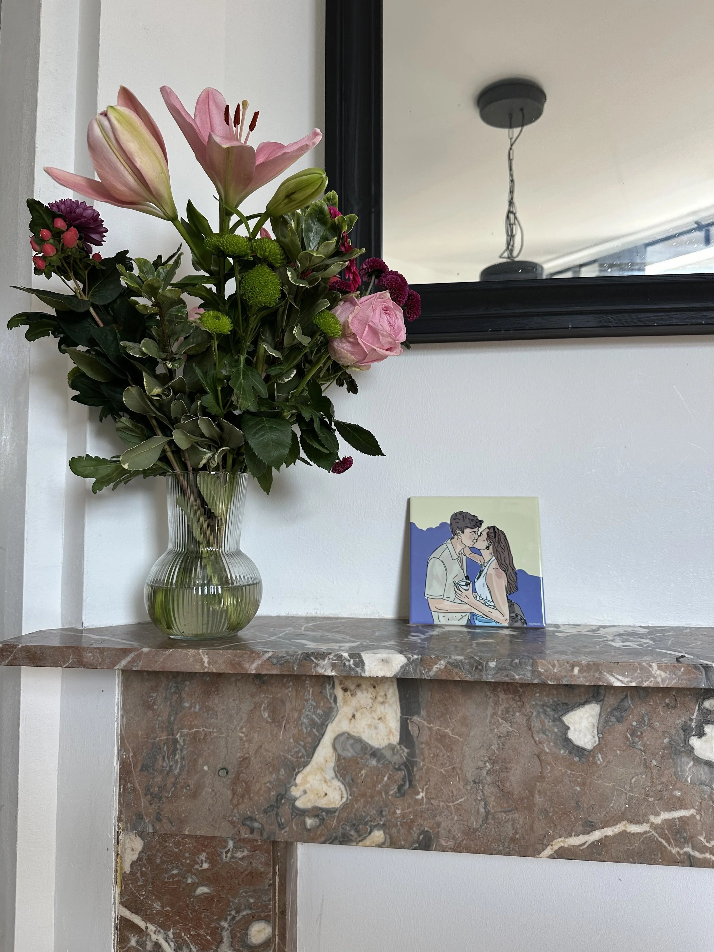 Een boeket bloemen in een glazen vaas naast een illustratie van een koppel dat elkaar kust, geplaatst op een marmeren blad, met een spiegel op de achtergrond.