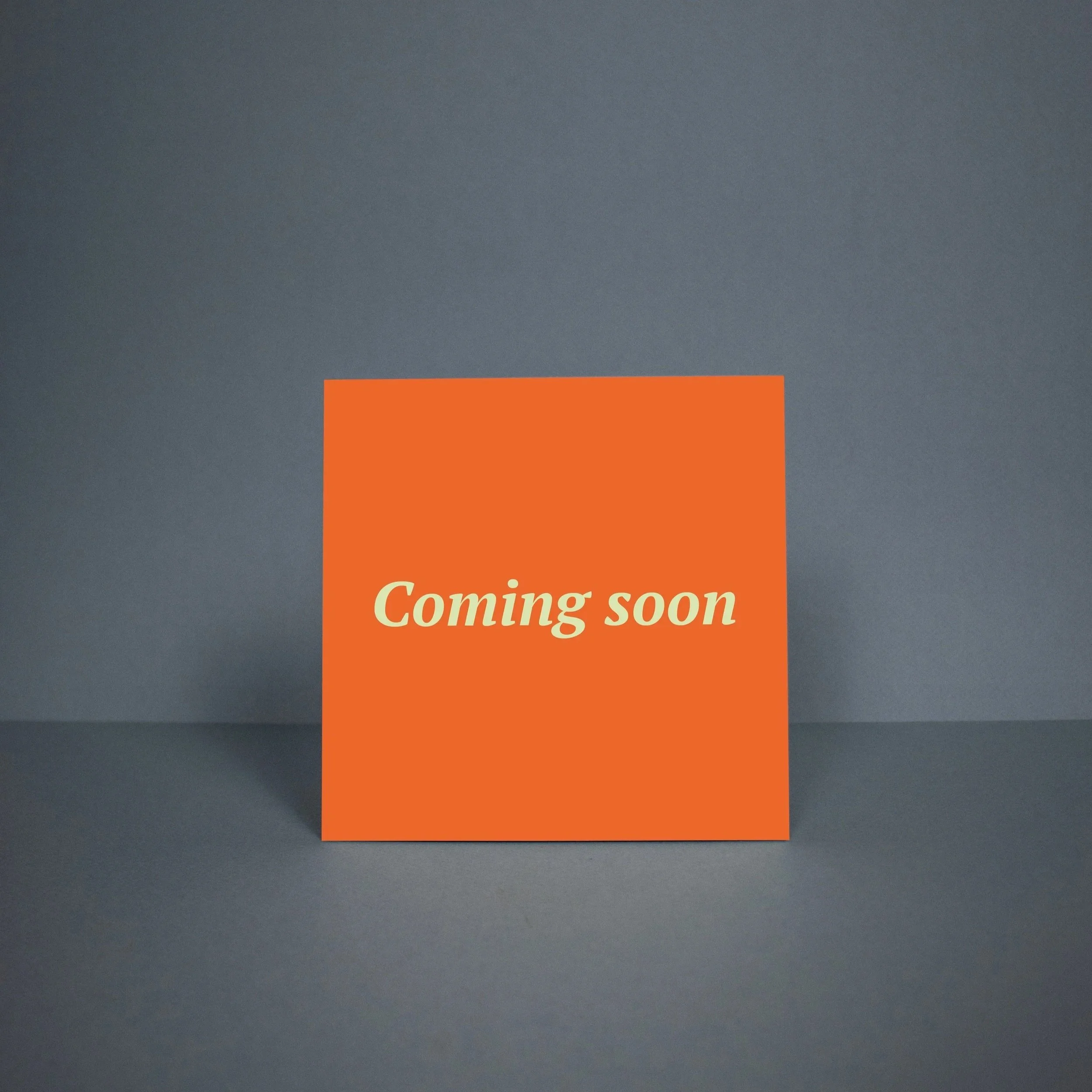 Een oranje vierkante bord met de tekst 'Coming soon' in het midden, geplaatst op een donkere ondergrond tegen een grijze muur.