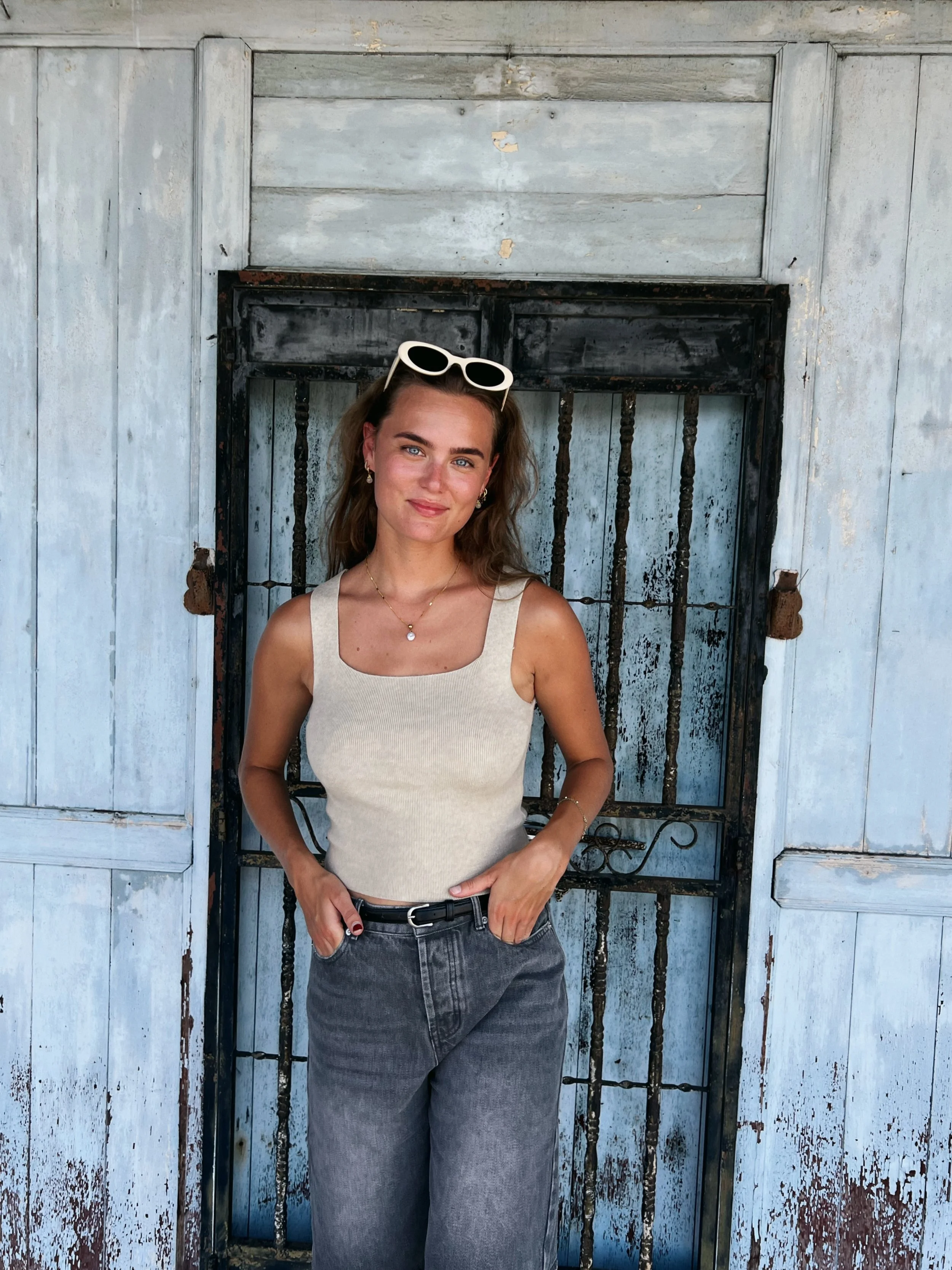 Een vrouw met bruin haar, zonnebril op haar hoofd, draagt een beige top en grijze jeans, staat voor een oude, verweerde houten en metallieke deur.