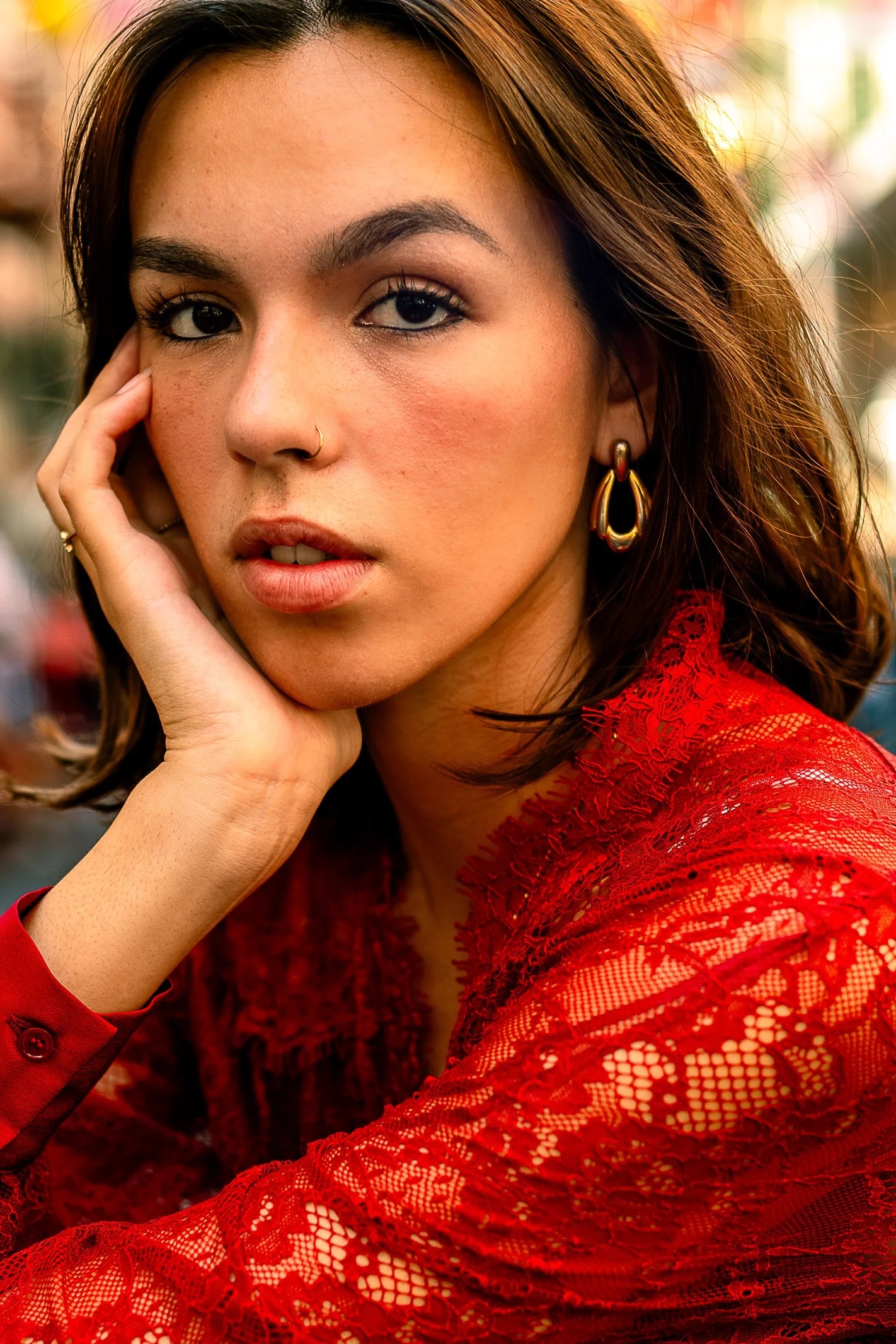 Close-up portrait of a young woman with brown hair wearing a red lace top, gold hoop earrings, and a nose ring. She is resting her face on her hand and looking at the camera with an outdoor background.