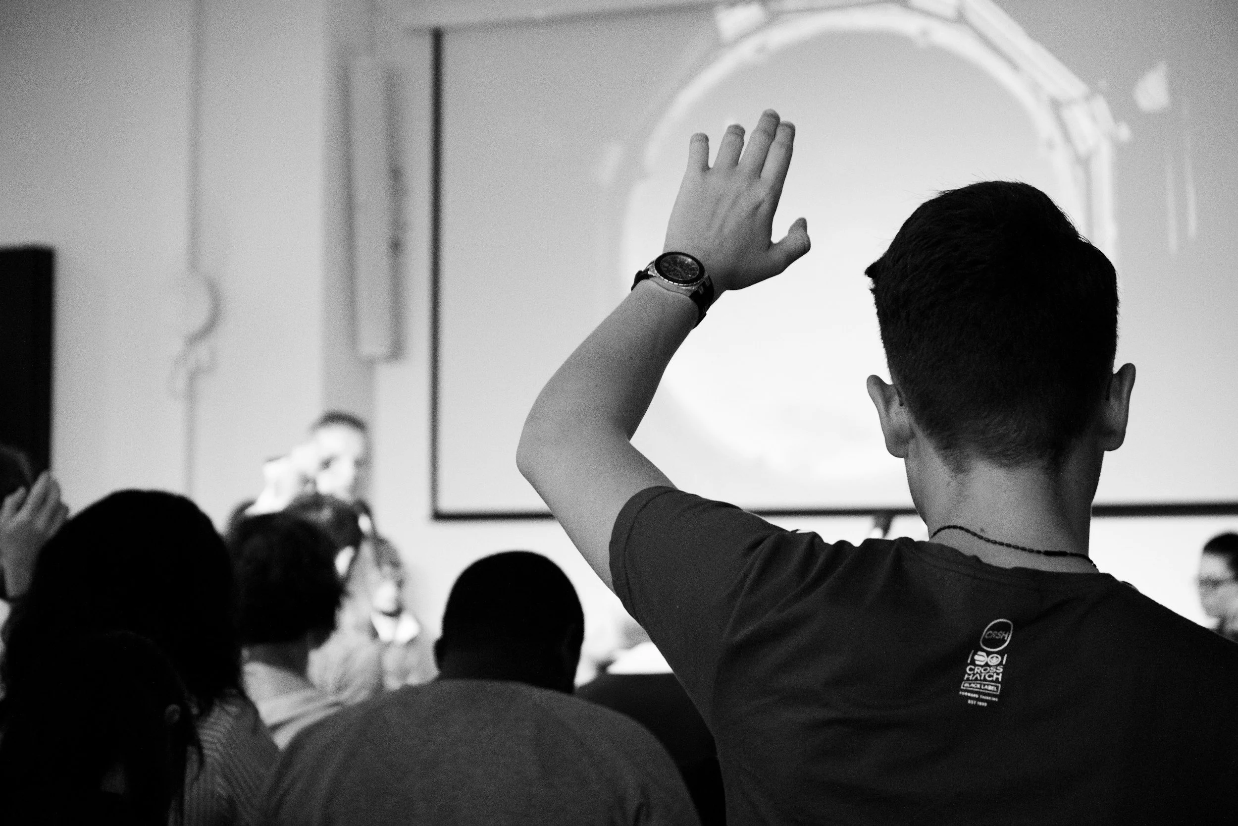 A person raising their hand during a presentation or lecture in a classroom, with other students visible in the audience, and a screen displaying an image or slide in the background.
