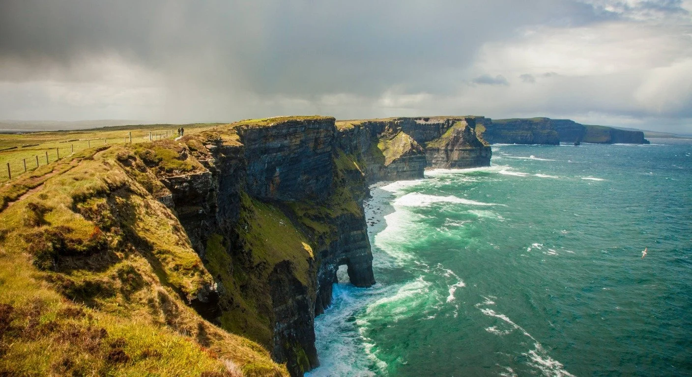 Dramatic steep dark cliffs meet the crashing white waves of the turquoise ocean under a moody, overcast sky with walking path on the left