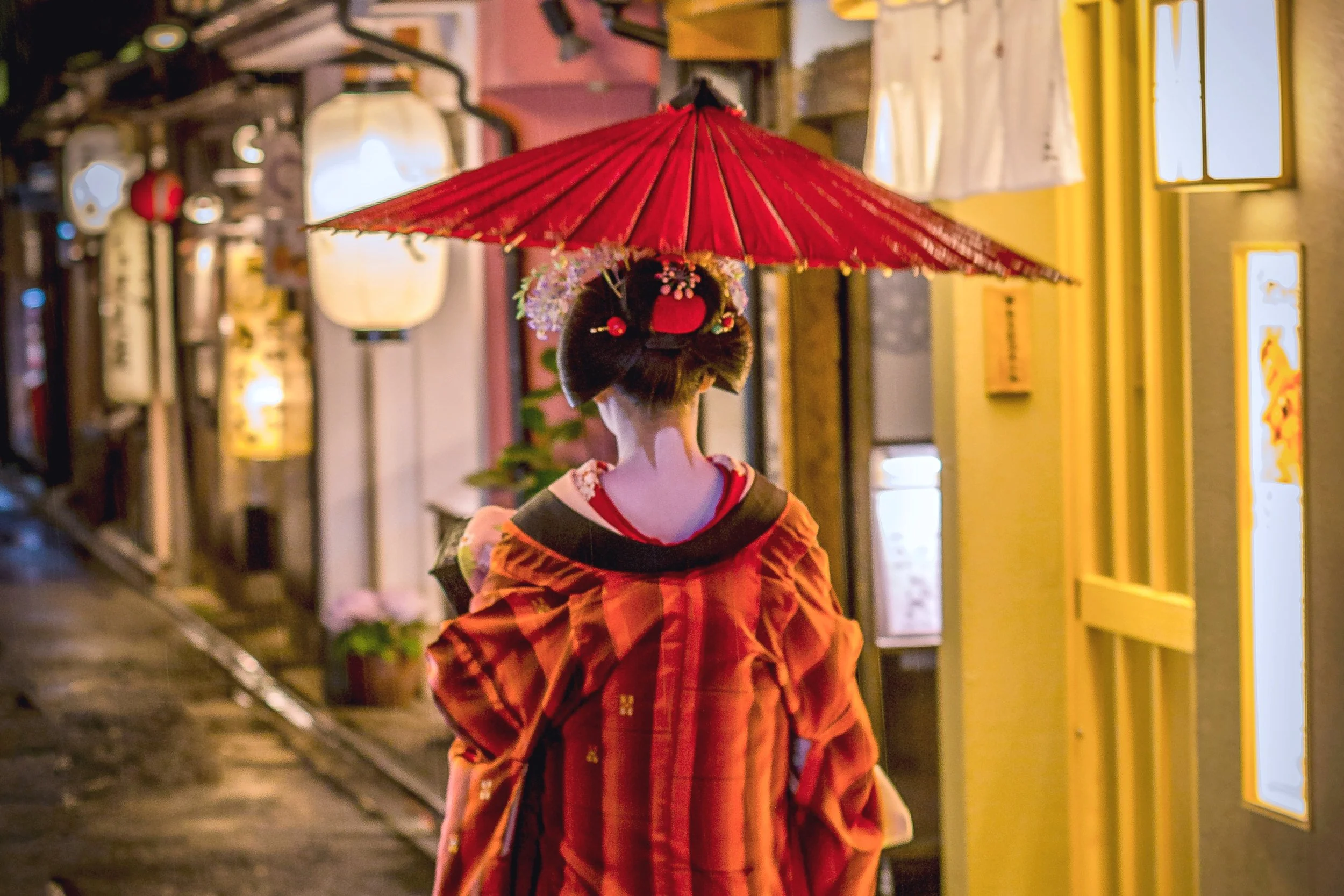 **Refined alt text (gallery image):**  Geisha walking through a lantern-lit Japanese alley, wearing a colorful kimono and carrying a red parasol, viewed from behind in a serene traditional setting.
