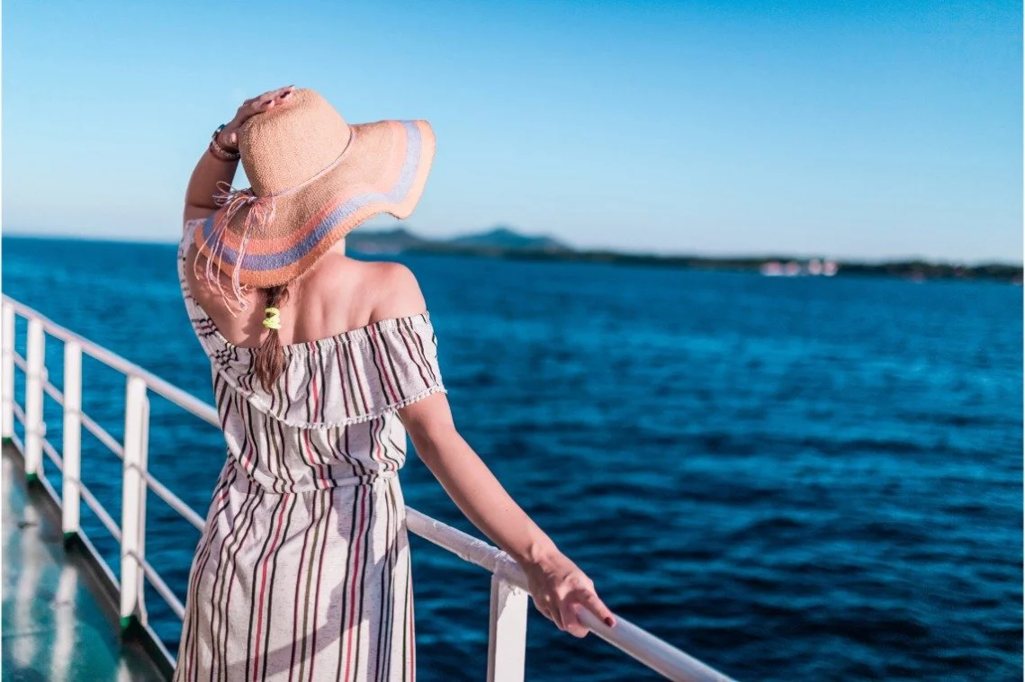 **Refined alt text (gallery image):**  Woman wearing a straw hat and striped dress standing on a boat, looking out over deep blue ocean under a clear sky, capturing a relaxed travel moment.