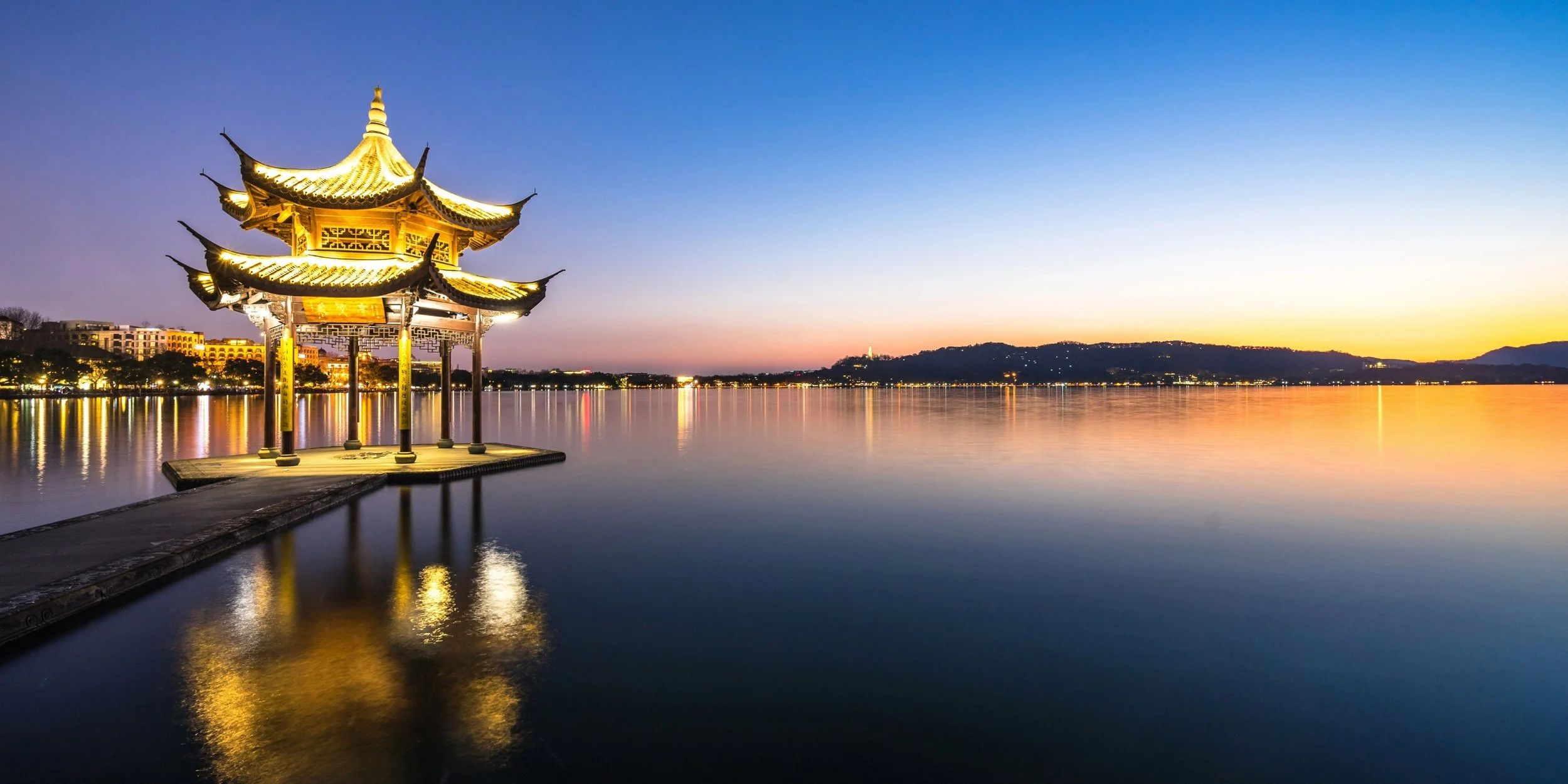 **Refined alt text (gallery image):**  Traditional Chinese pavilion glowing at twilight over calm water, with golden lights reflecting beneath a blue-to-orange sky and distant mountains silhouetted on the horizon.