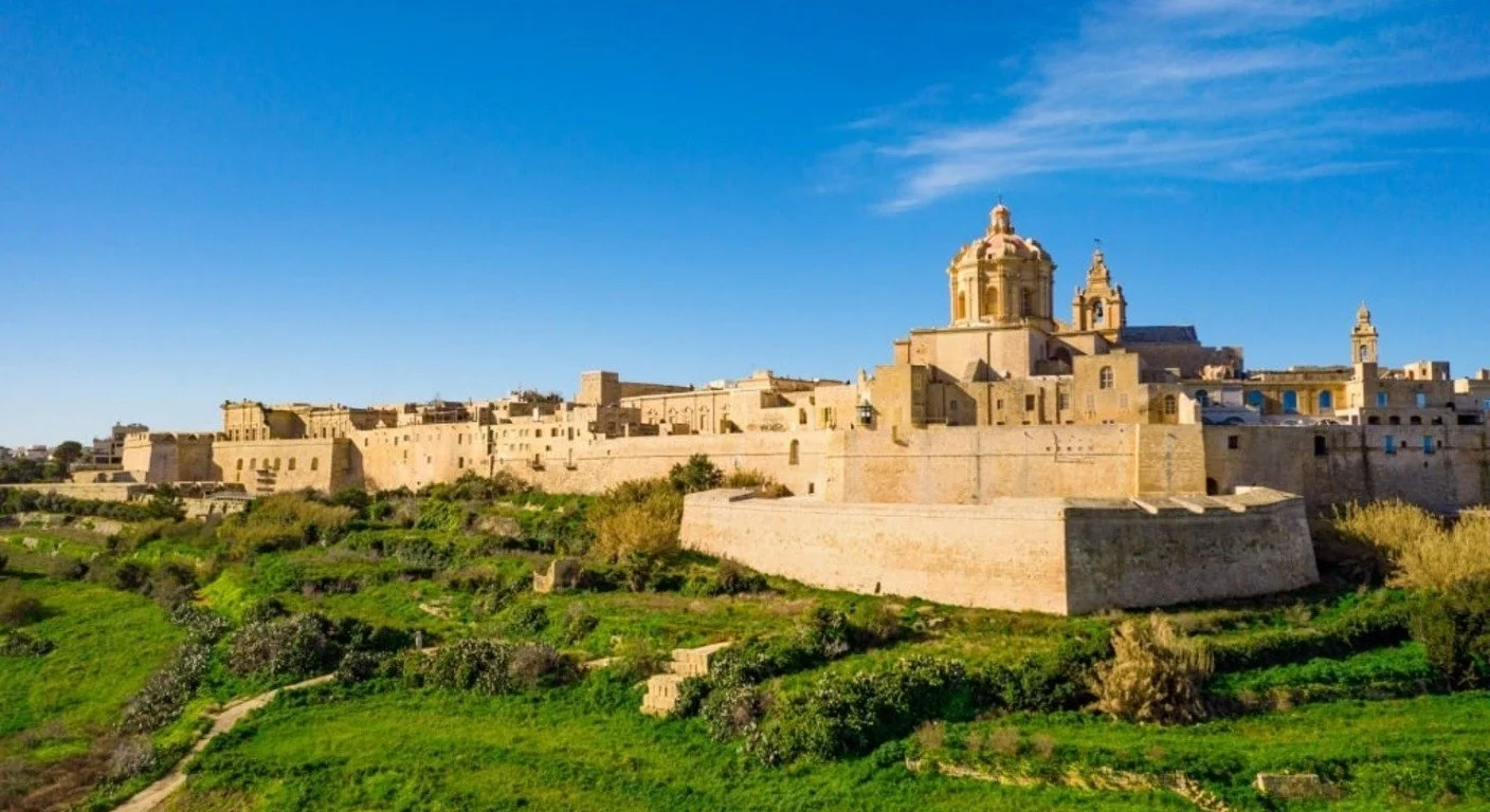 A wide panoramic view of the fortified limestone city of Mdina on a hill, surrounded by green fields and trees under a clear blue sky.