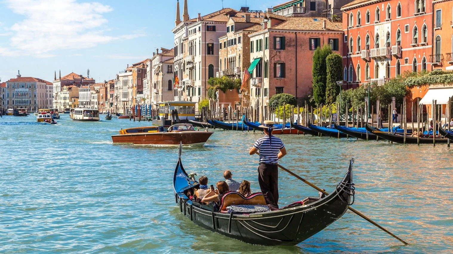 Gondola with passengers gliding along Venice’s Grand Canal, guided by a gondolier, with sunlit historic buildings and turquoise water in a lively city scene.