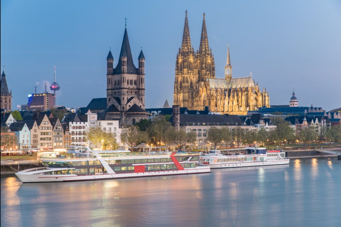 **Refined alt text (gallery image):**  Cologne cityscape at dusk with Cologne Cathedral spires and St. Martin Church glowing beside the Rhine, reflecting warm lights on calm river water.