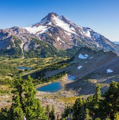 Snow-capped mountain with green foothills, small lakes, and trees in foreground under a clear blue sky.