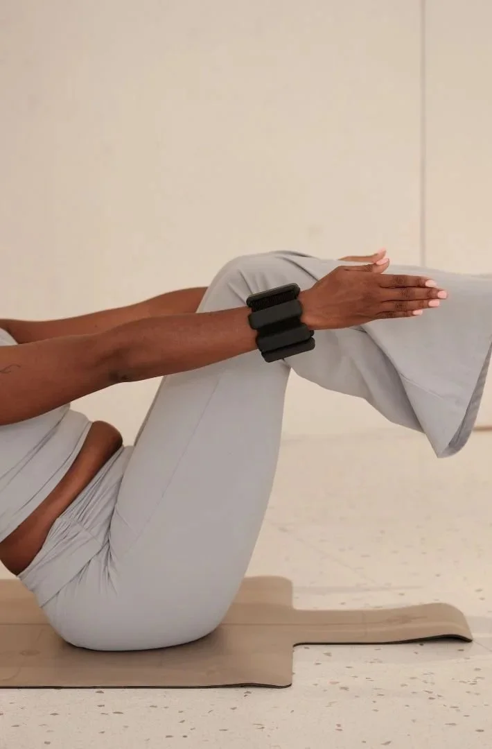 Personne en posture de yoga avec les jambes pliées, tenant une jambe levée avec les mains, portant une montre ou un bracelet. Assise sur un tapis beige dans une pièce intérieure.