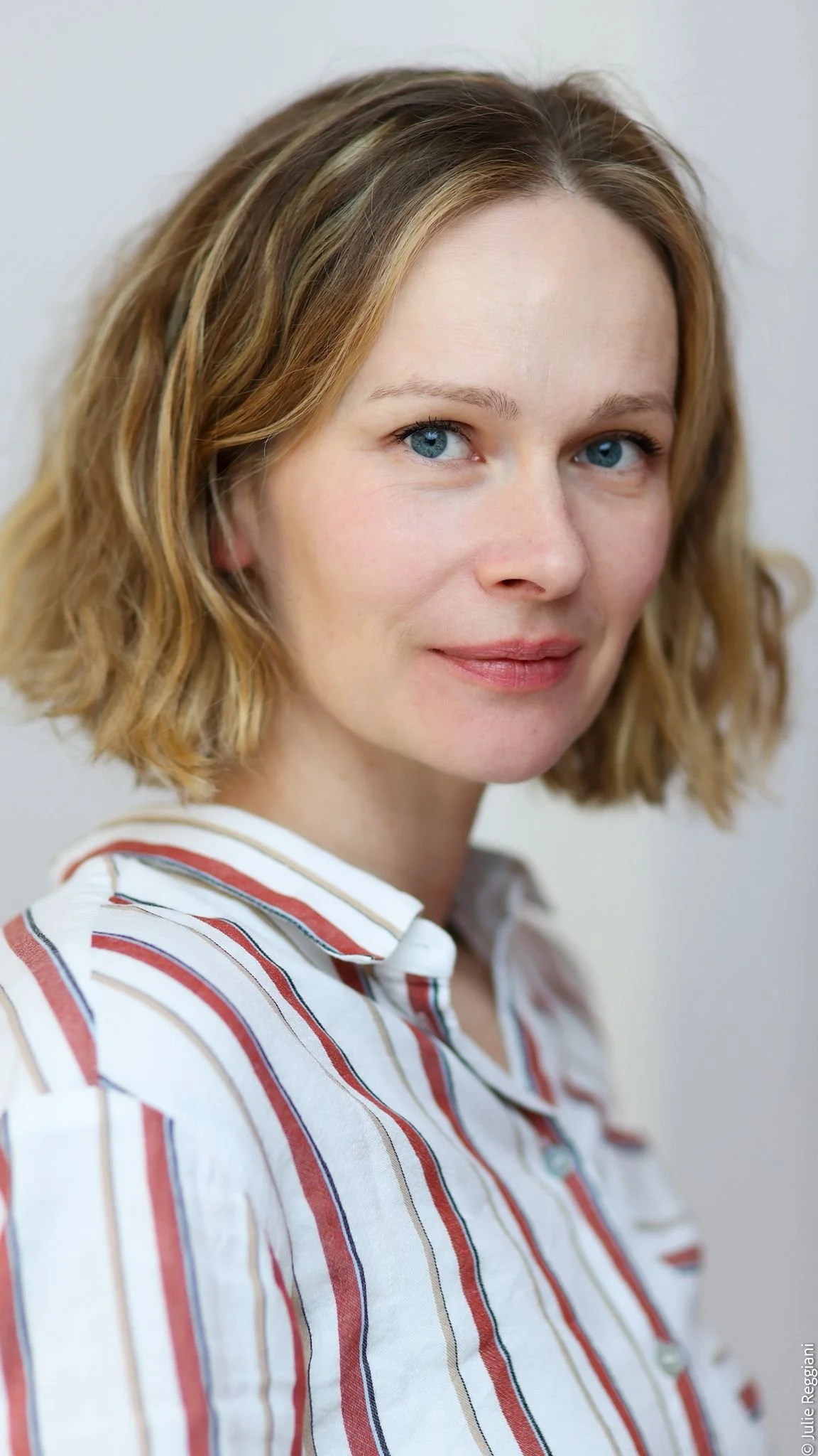 Portrait d'une femme aux cheveux courts, aux yeux bleus, portant une chemise à rayures rouges, blanches et noires.