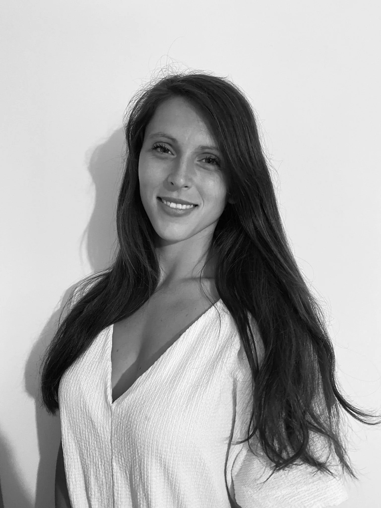 Portrait en noir et blanc d'une femme avec de longs cheveux ondulés, portant un vêtement blanc, souriant légèrement, avec un mur blanc en arrière-plan.