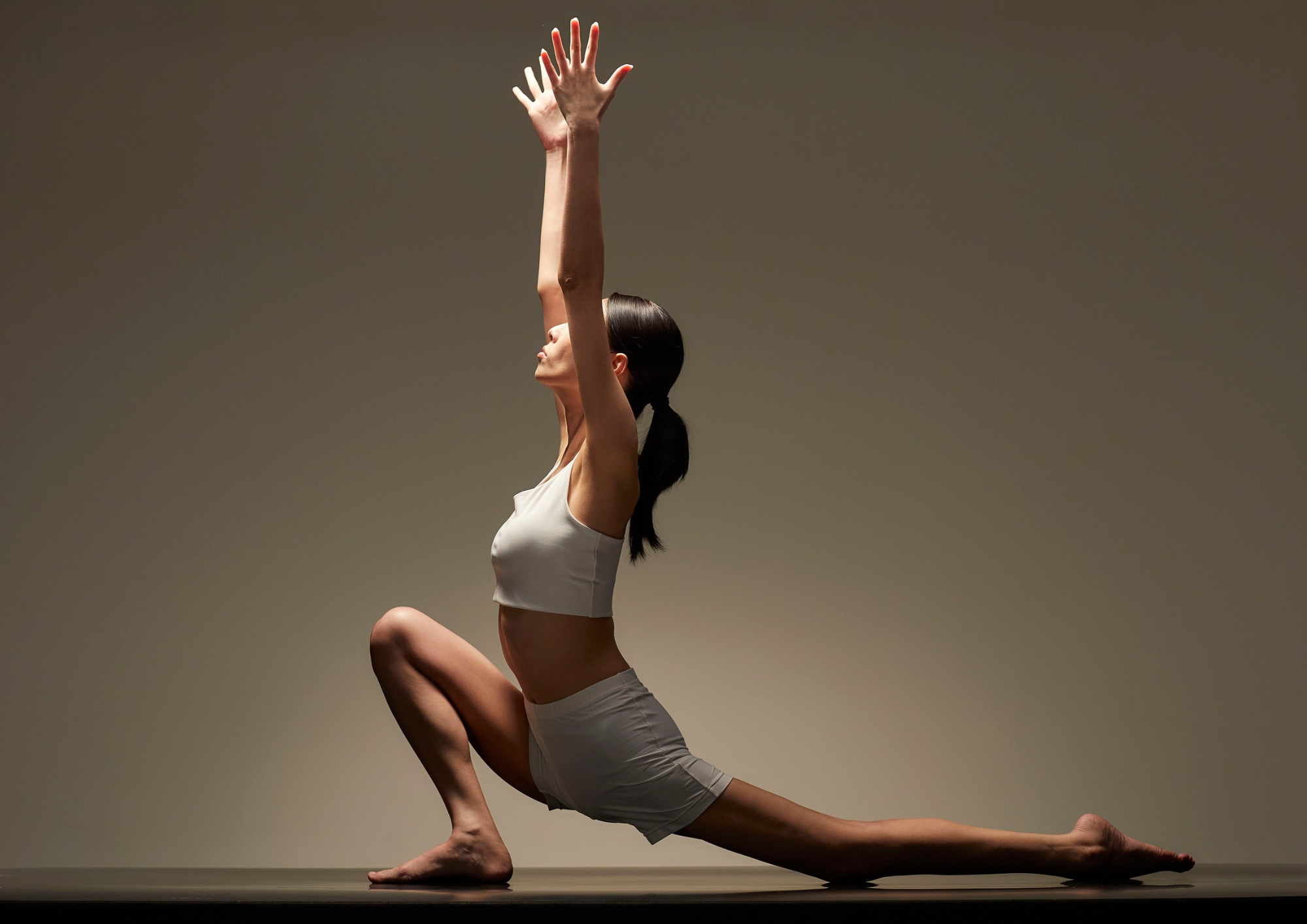 Une femme pratique le yoga en position de guerrier sur un sol noir, dans une pièce sans décor avec un fond beige uniforme.