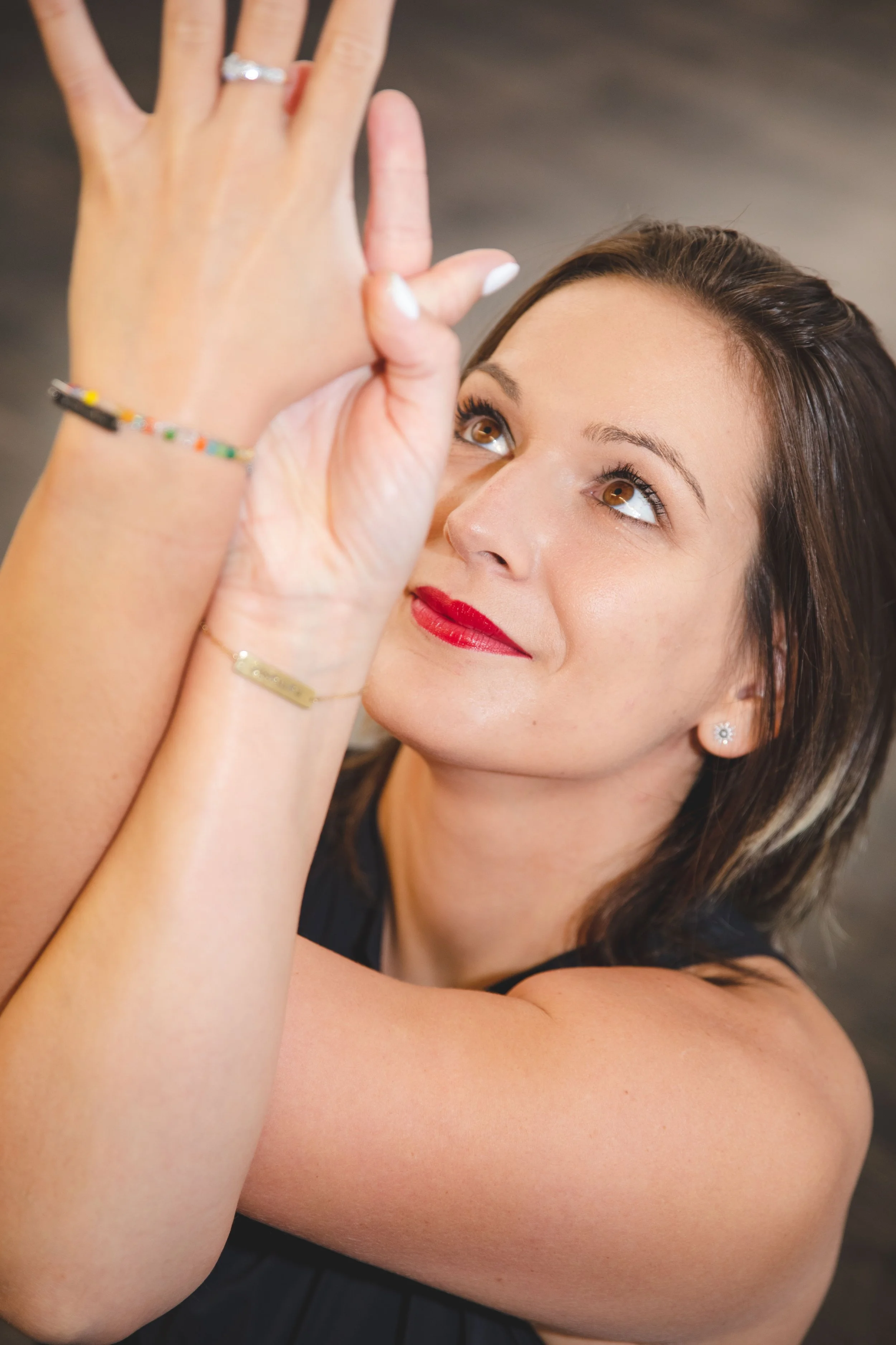Une femme souriante avec des cheveux bruns et des yeux marron, portant du maquillage rouge à lèvres, qui fait un geste avec sa main et porte des bijoux, notamment une bague et un bracelet, dans un cadre intérieur.