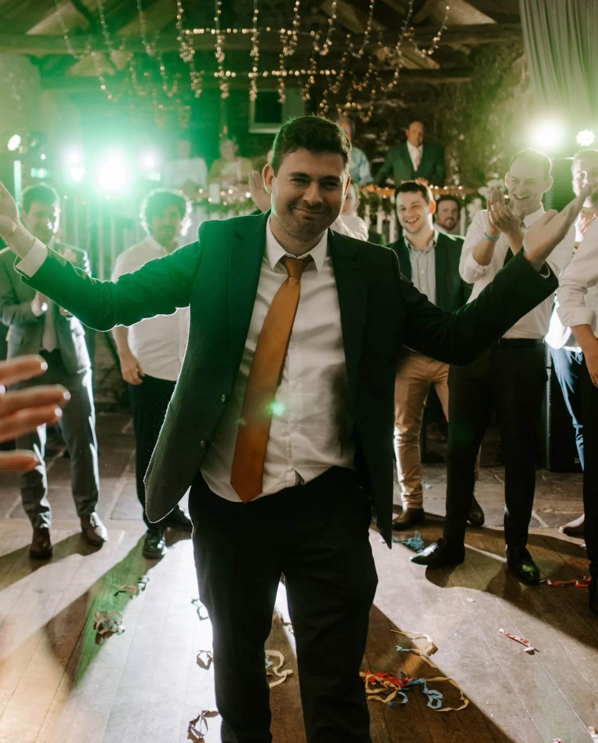 Man in black suit with orange tie dancing at a celebration party, with others clapping and smiling in the background. Bright lights and streamers are visible.