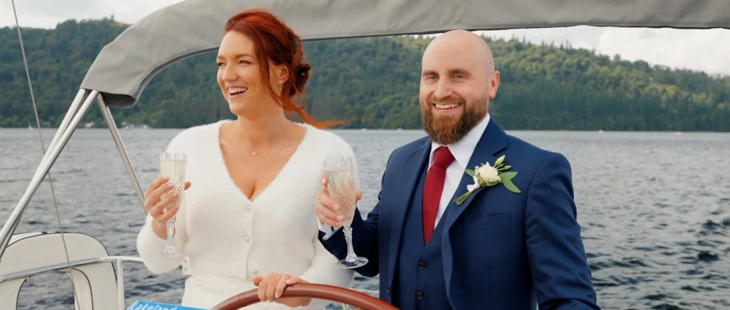 Smiling newlyweds on a boat holding champagne glasses on Lake Windermere.