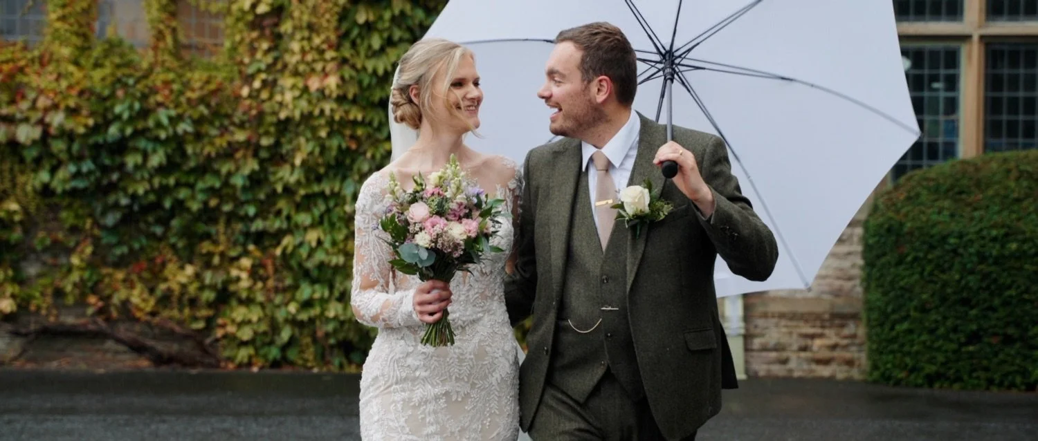 A bride and groom walking outside in the rain, sharing a joyful moment under an umbrella. The bride is holding a bouquet and wearing a lace wedding dress. The groom is dressed in a suit with a boutonniere.