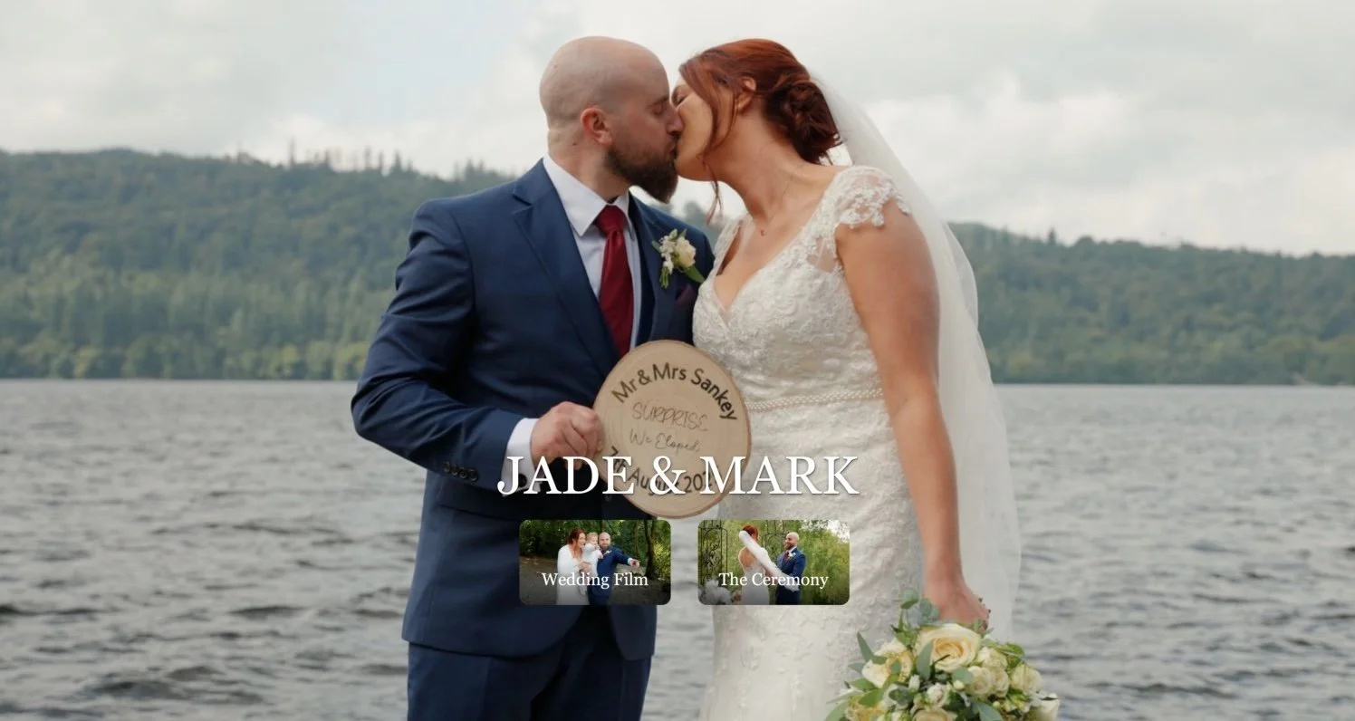 A newlywed couple, Jade and Mark, kiss by a Windermere with a green forested hillside in the background, after their wedding ceremony.