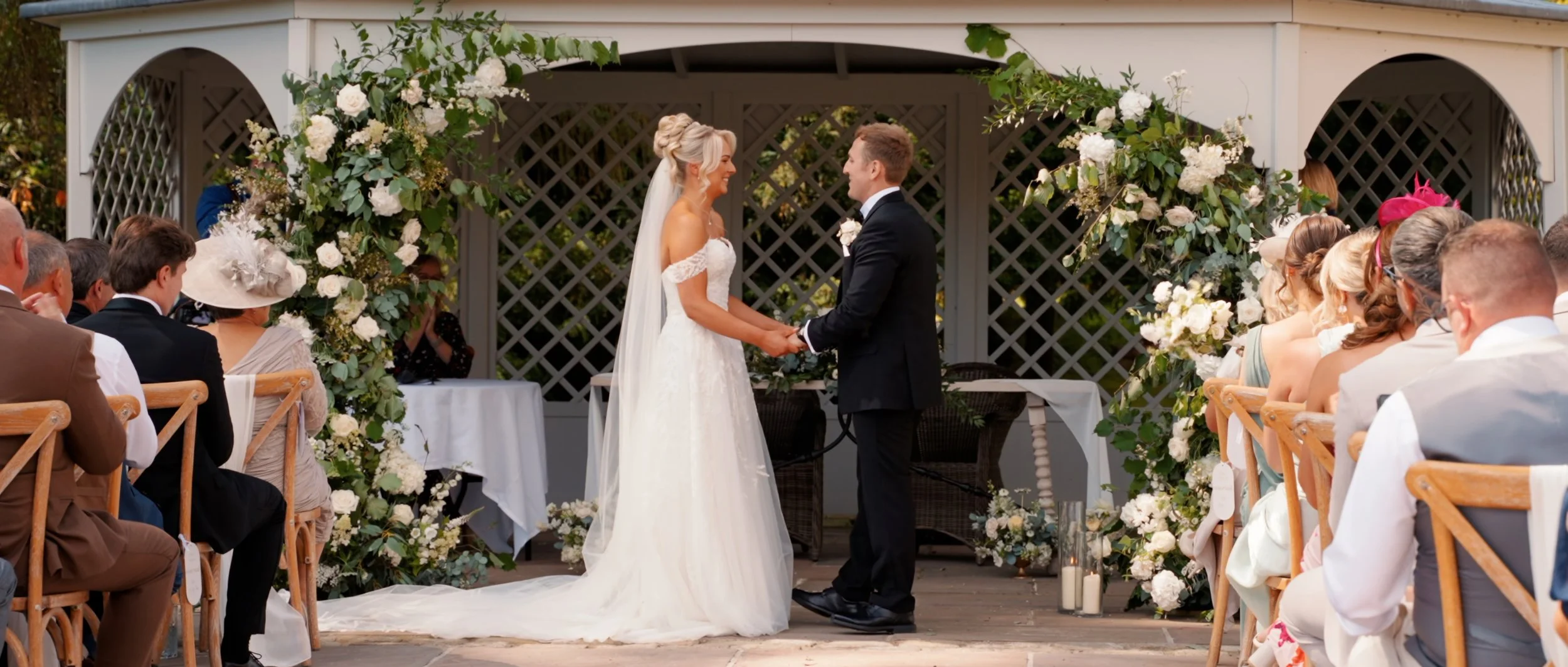 Bride and groom holding hands during outdoor wedding ceremony surrounded by guests and floral decorations.