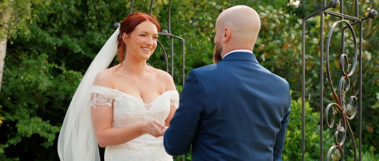 A bride and groom holding hands and exchanging vows outdoors, with greenery in the background in the Lake District.