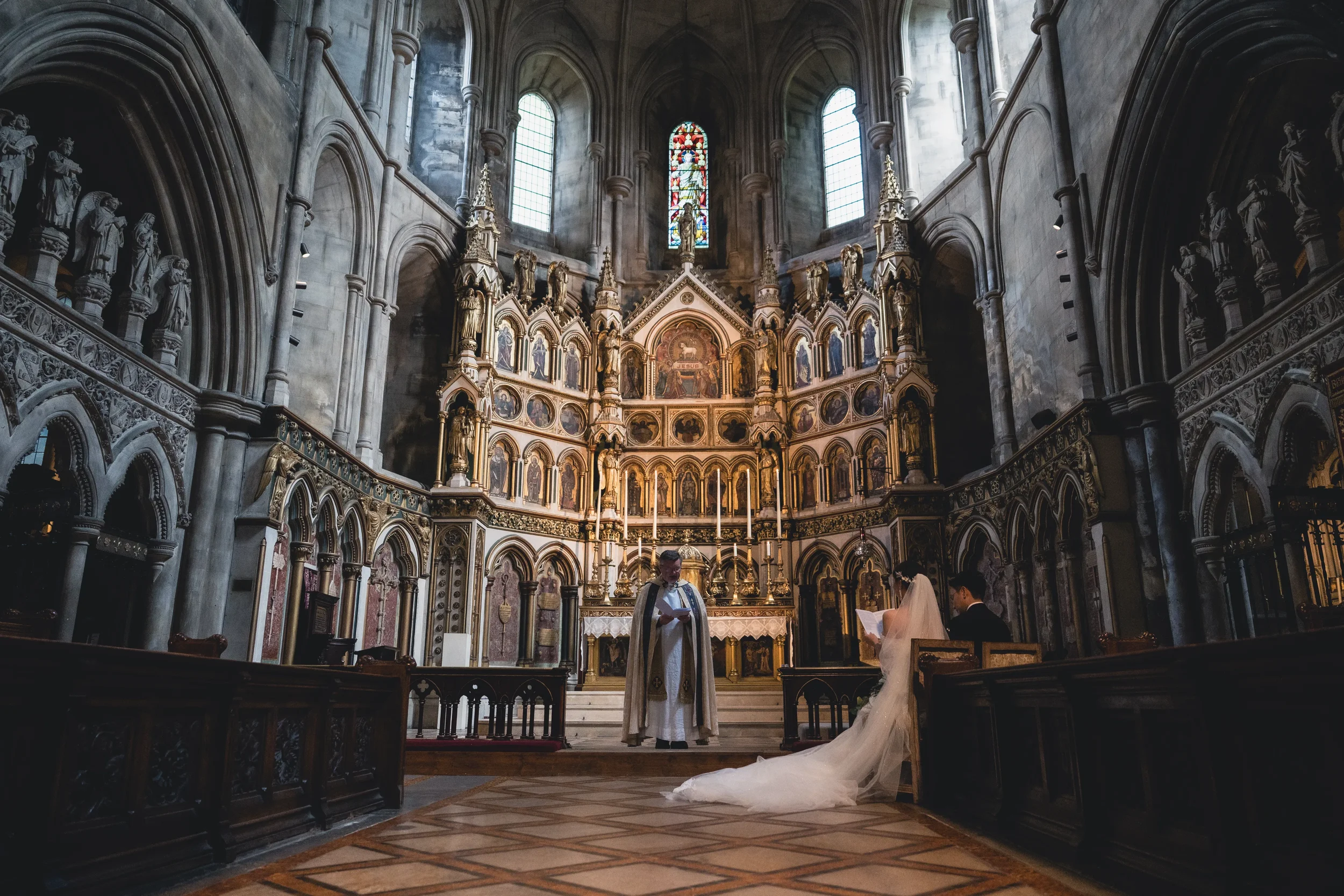 教会での結婚式の場面。中央に司祭と新郎新婦が立っており、新婦は白いドレスを着ている。背景には装飾された祭壇とステンドグラスの窓が見える。