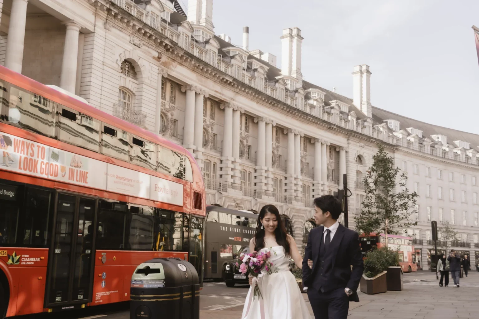 結婚式の衣装を着た女性と男性が街中を歩いている。背景には歴史的な建物と赤い二階建てバスが見える。