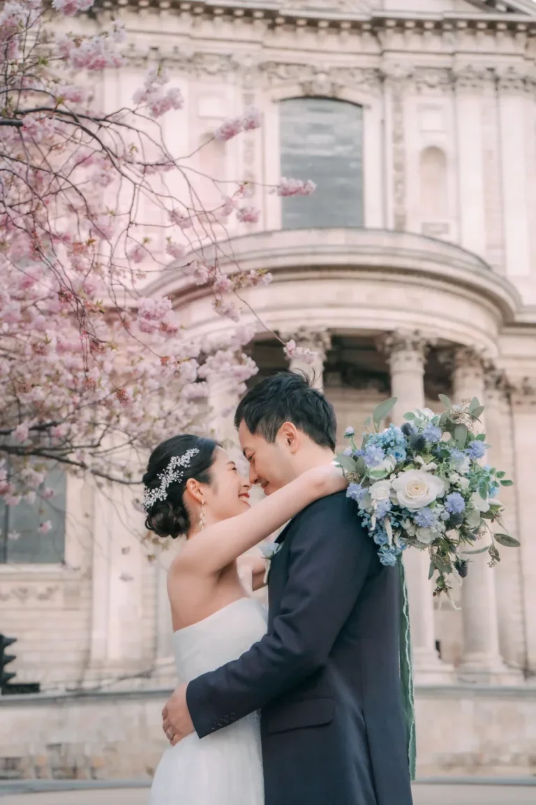 桜の木の下で抱き合う新郎新婦。背景に歴史的な建物。