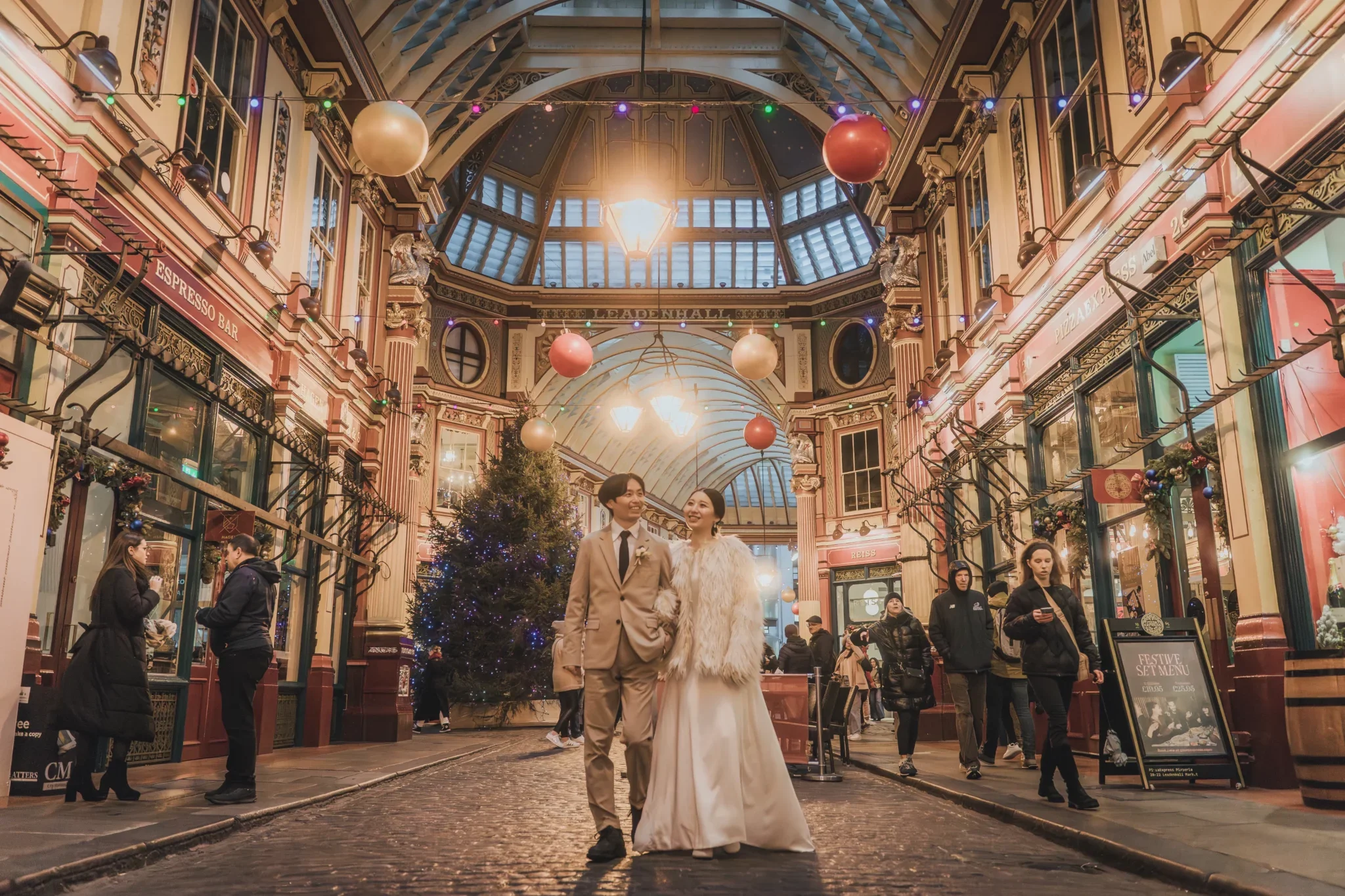 クリスマスの装飾が施されたショッピングアーケードの中央を歩く若いカップル。男性はベージュのスーツ、女性は白いドレスとファーコートを着ている。周囲にはクリスマスツリーや飾り付け、買い物客と看板が見える。
