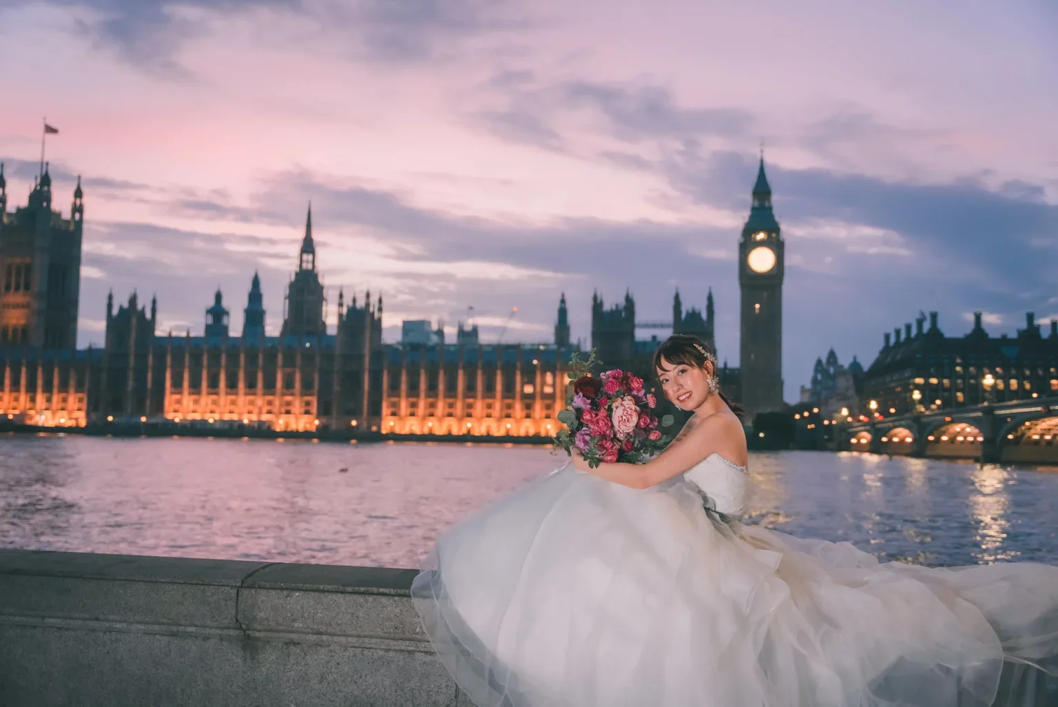 結婚式のドレスを着た女性が川辺で花束を持ち、夜のロンドンの風景を背景に微笑んでいる