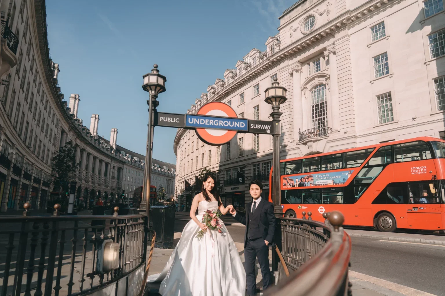 ロンドンの地下鉄入り口前で、新郎新婦が並んでいる。背景には赤い二階建てバスと歴史的な建物が見える。