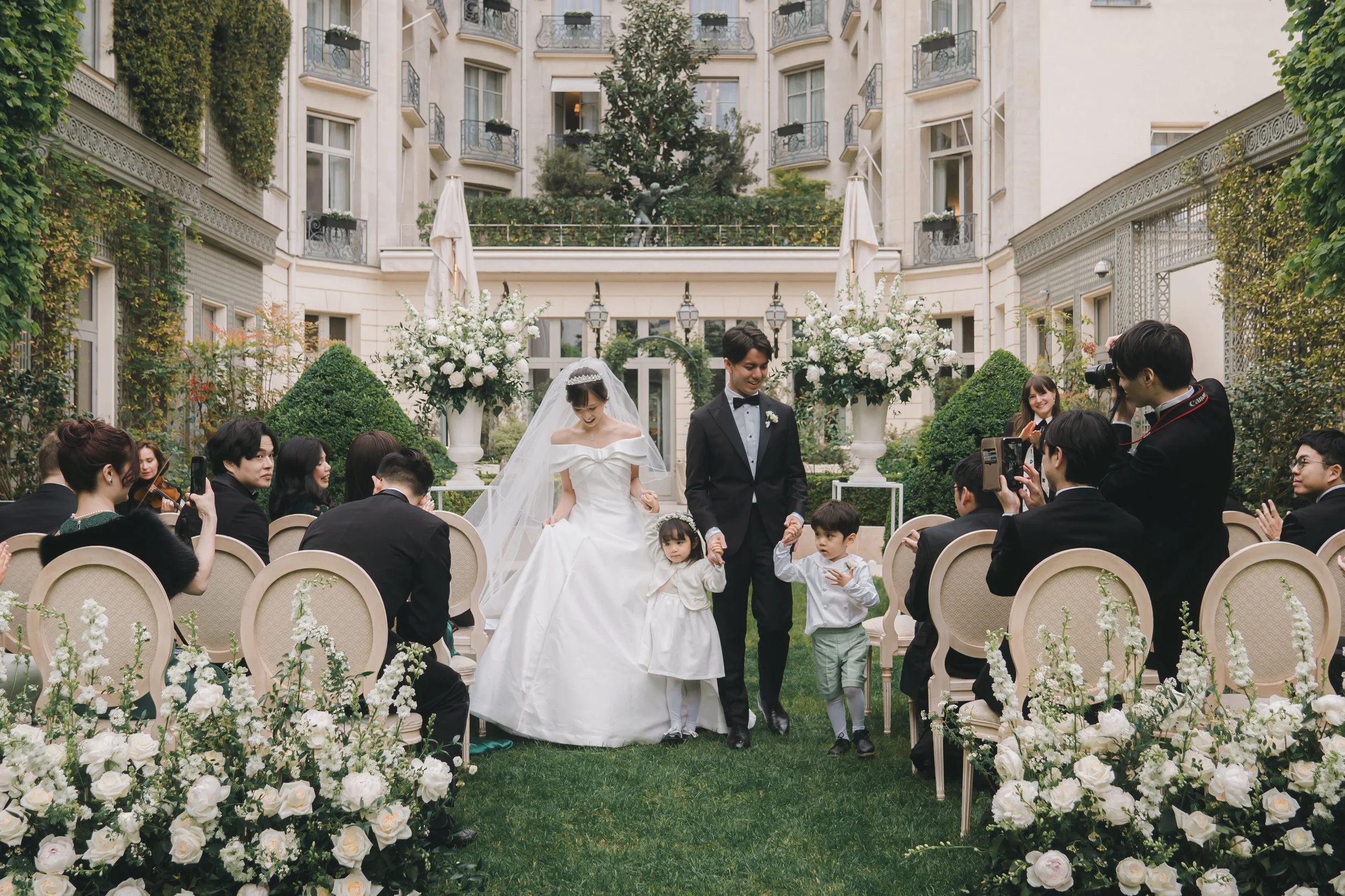 結婚式のセレモニーで新郎新婦が子供たちと共に歩く様子、ゲストとカメラマンが見守る庭園のロケーション
