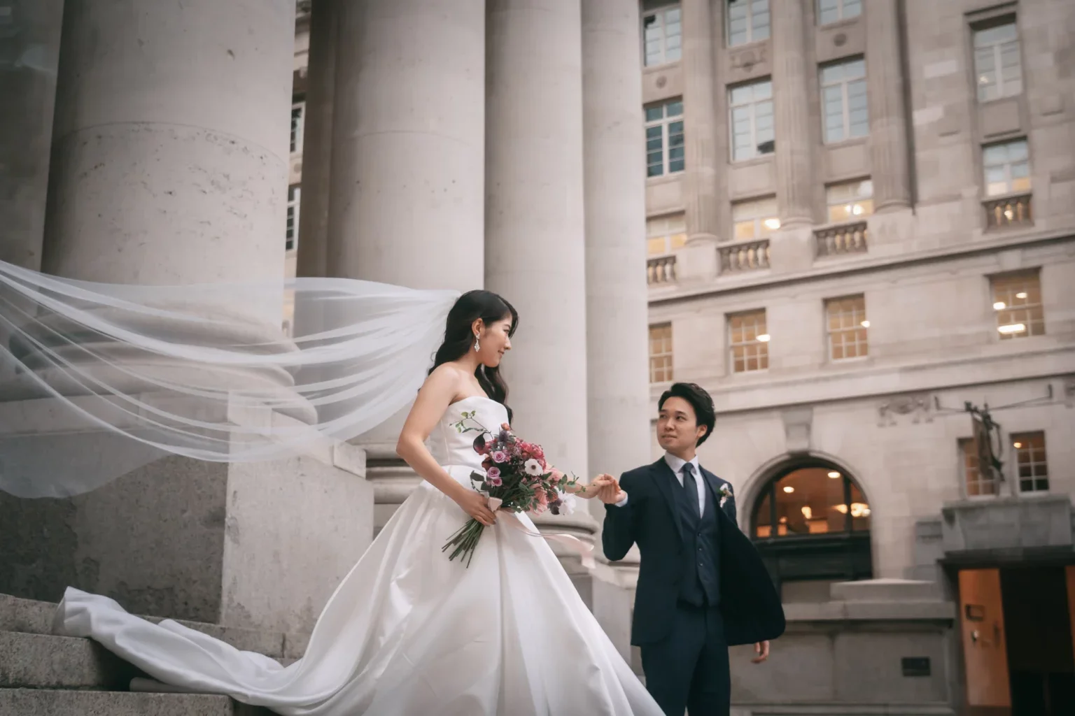結婚式の新郎と新婦が階段の上で手を取り合う。新婦は白いドレスを着て花束を持ち、長いヴェールが風になびいている。背景は古典的な建物の外観。