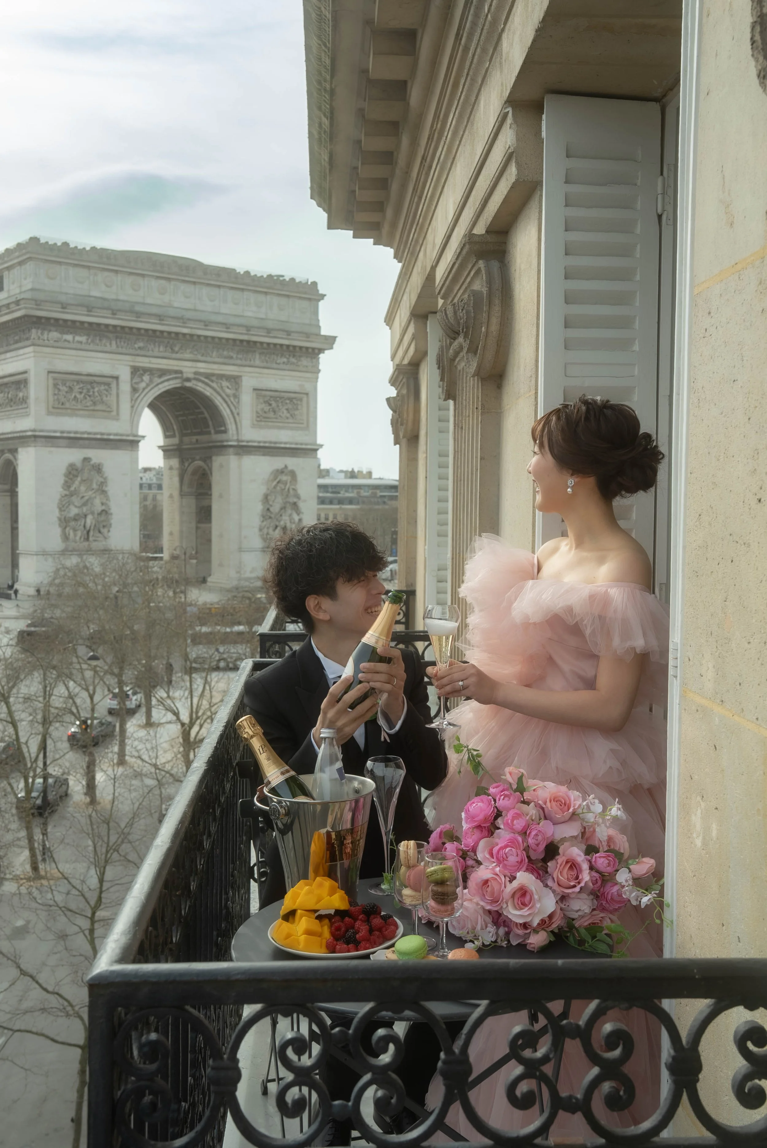 パリのエッフェル塔を背景にしたバルコニーで、結婚式を祝うカップルがシャンパンを交換しながら笑顔で見つめ合っている。テーブルにはフルーツやケーキ、シャンパンが置かれている。」}}