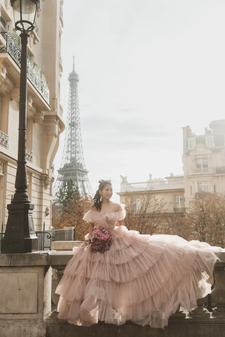 パリの街角で、ベルサイユ風の豪華なピンクのドレスを着た女性が花束を持ち、背景にエッフェル塔が見える風景