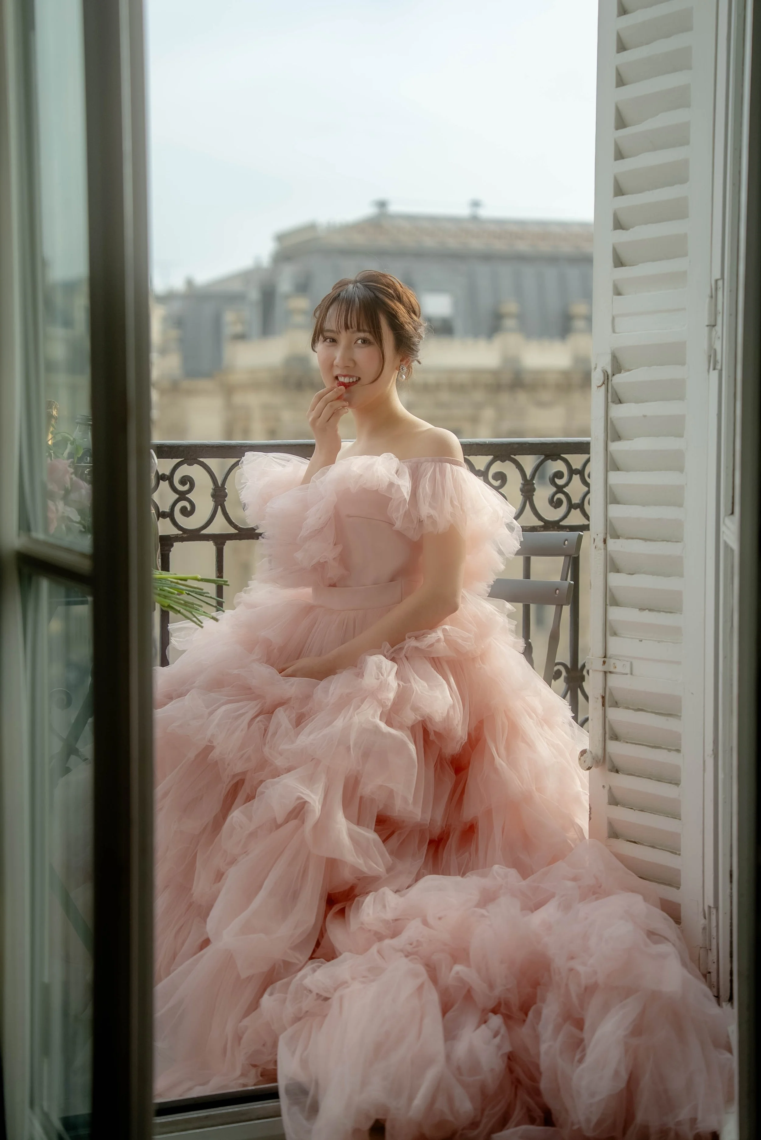 ピンク色のふわふわとしたドレスを着た若い女性がバルコニーで微笑みながら指を唇にあてている。背景には古い建物の屋根が見える。