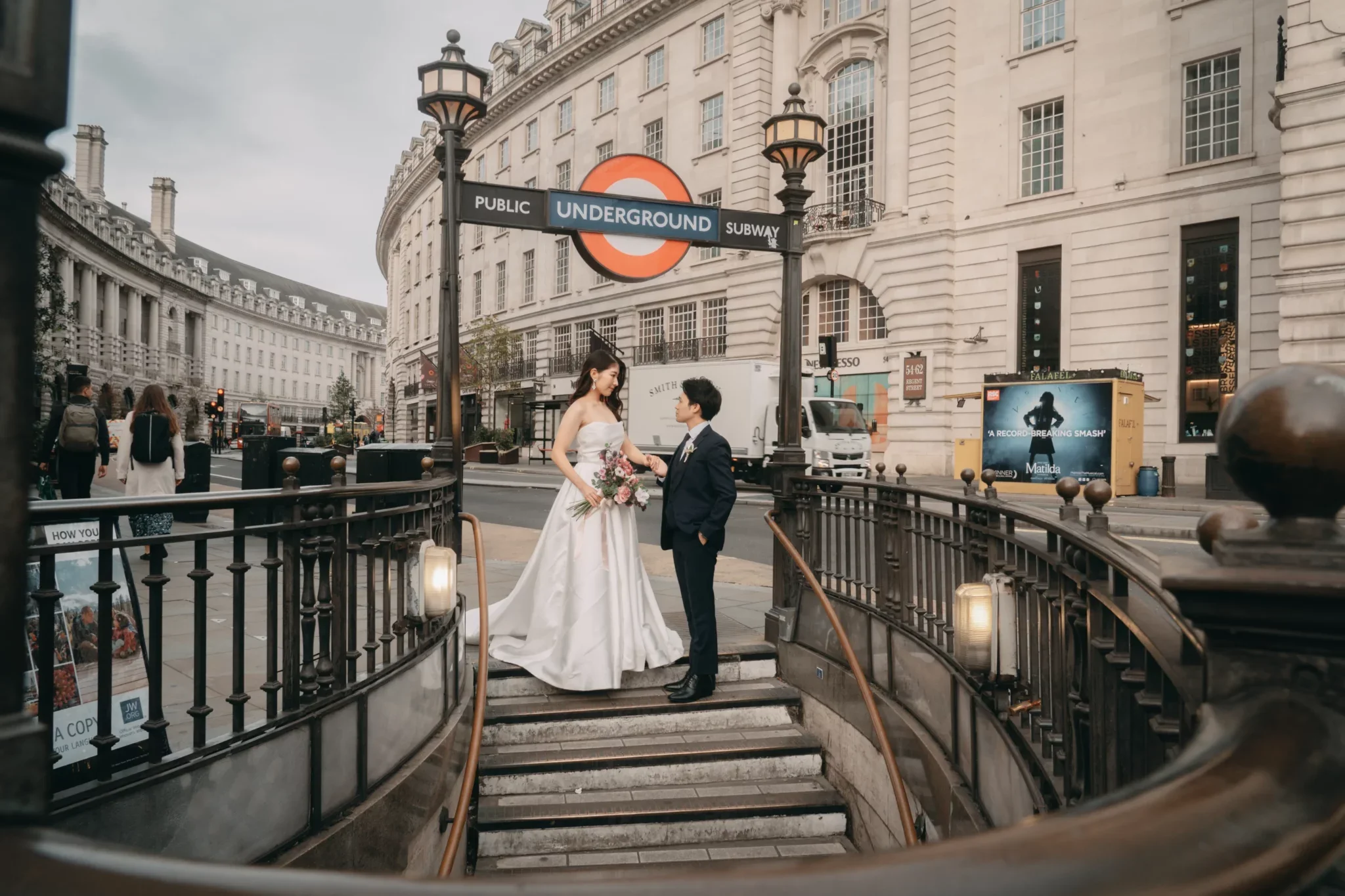 ロンドンの地下鉄入り口前で、結婚式を挙げているカップル。女性は白いドレスと花束を持ち、男性はスーツを着ている。