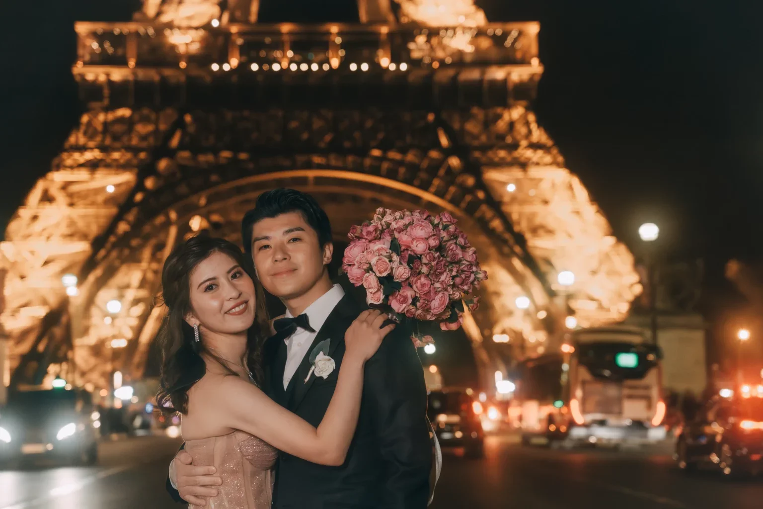 夜のパリ、エッフェル塔の前で結婚式を迎えた新郎新婦が抱き合って笑顔を見せている。新婦はピンクのドレスを着て、花束を持っている。背景には輝くエッフェル塔と交通量の多い道路が見える。
