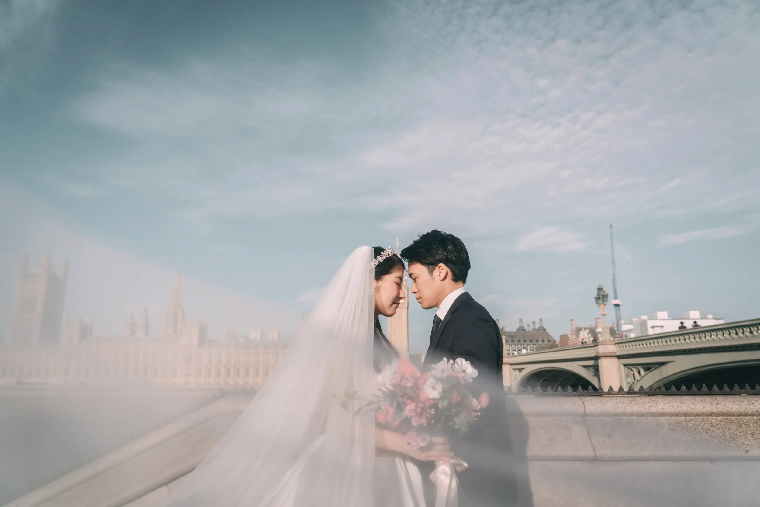 結婚式を挙げているカップルが顔を近づけている。背景にロンドンのビッグベンと橋が見える晴れた空の下の都市の景色。